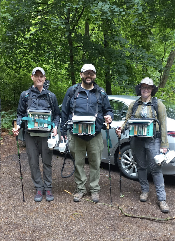 Three researchers with LI-6800 consoles strapped in, holding a walking stick in one hand and the LI-6800 console head in another. Silver car and trees apparent in background